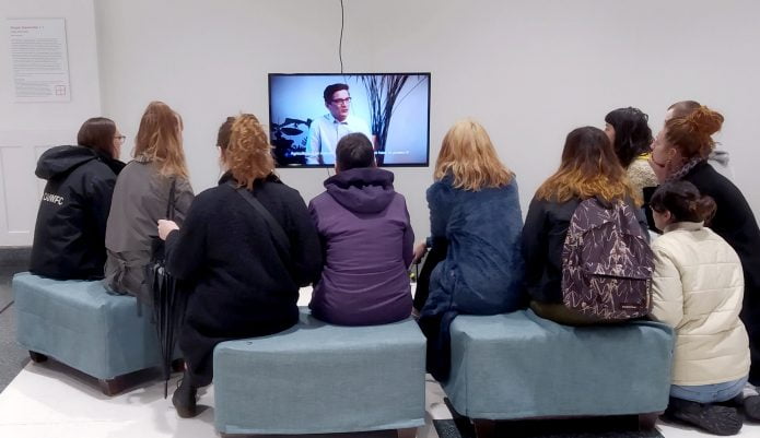 A group of Level two DJCAD students sit on grey cubes in a white room, watching a video artwork on a small screen. The video artwork is Utopia Generator by artist Julia Schicker, and the screen shows a white person in glasses in mid-sentence.