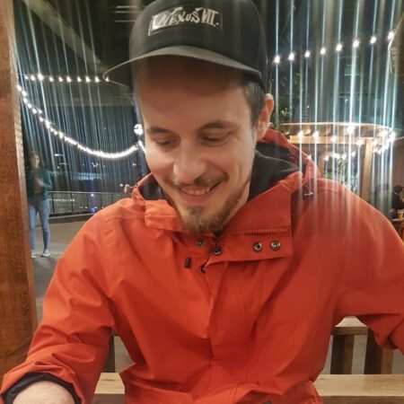 Picture of a man wearing cap and orange wind breaker jacket who is smiling and looking down.