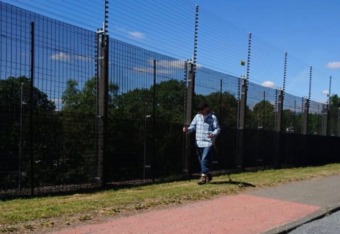B.D. Owens walks alongside the fence at Faslane naval base, marking the #UndesiredLine with his modified shoes and hiking sticks.