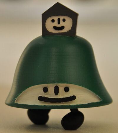 The first prototype toy figure of Akinori Oishi's LAWHILL – 3D design by Creo Design from Dundee city (2010) . The figure is green in colour with a white face. The shape of the figure is in the form of a hill. On the top of the figure is a grey houselike shape, also with a white smiling face.