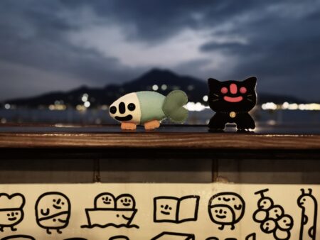 A homemade green and white fish plush toy and a homemade black cat plush toy standing on a window ledge.