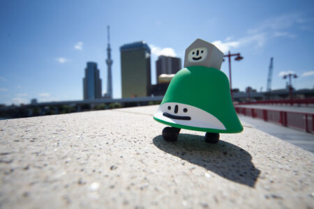 The Law Hill in Dundee depicted in toy form by Japanese artist Akinori Oishi. The toy is green and hill shaped, with a white smiling face. On top of the hill is a small grey houselike shape also with a white smiling face. The figure is sitting on a wall. In the background are large office blocks.