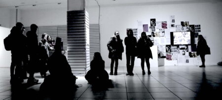Exhibition Audience. A black and white image of a group of people in a white room looking at images on a wall.