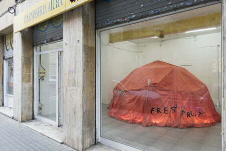 Installation by Holly White. Image shows artwork through a shop window. Artwork comprises a large orange tent with the words Fresh Fruit written in black letters on the side of the tent.