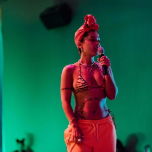 Image of Fannie Sosa singing Fannie is coloured orange, wearing a tiger skinned bikini top and orange jogging bottoms, singing into a microphone in a room thats green.