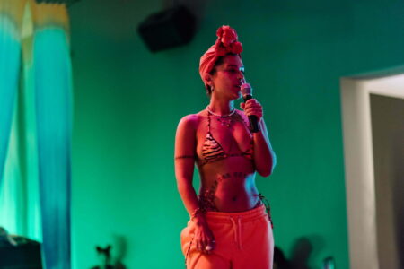 Image of Fannie Sosa singing Fannie is coloured orange, wearing a tiger skinned bikini top and orange jogging bottoms, singing into a microphone in a room thats green.