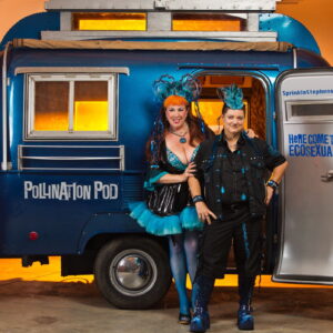 Annie Sprinkles and Beth Stephens in a room filled with orange light. They are standing in front of a small blue caravan with the words " Pollination Pod" written in white letters on the side. Written on the inside of the open door are the words " Here Come The Ecosexuals! " written in blue writing.