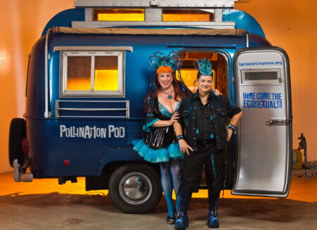 Annie Sprinkles and Beth Stephens in a room filled with orange light. They are standing in front of a small blue caravan with the words " Pollination Pod" written in white letters on the side. Written on the inside of the open door are the words " Here Come The Ecosexuals! " written in blue writing.