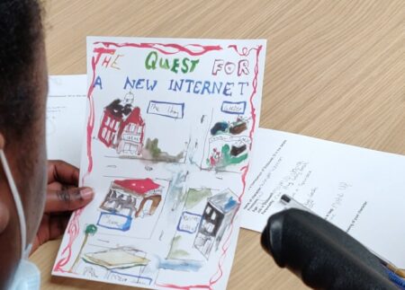 a young person sitting at a desk, holding a piece of paper with four illustrations on it. The heading on the paper reads " The quest for a new internet."