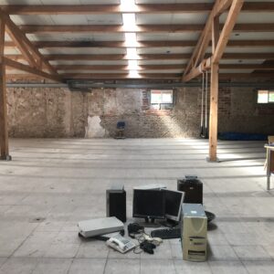 A large warehouse type room with old computers and old tech on the floor. Table and chair in background.