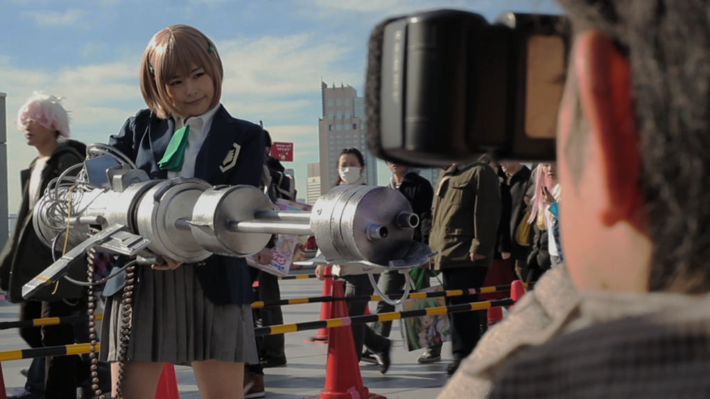 A Japanese schoolgirl holding a large , futuristic looking gun while a group of people look on.