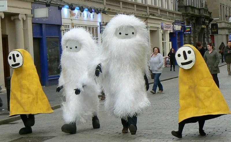 Four people dressed in costumes walking down a busy high street.Two people are dressed in white furry suits with grey faces. Either side of them is a person dressed in a yellow cover with no arms and a white face.