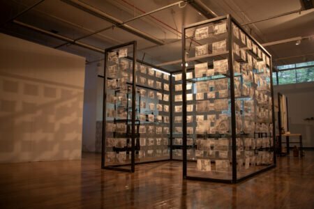 A black metal framed cube filled with rows of black and white images, printed on very thin paper.The cubed frame is situated in a white walled exhibition space with a wooden floor.